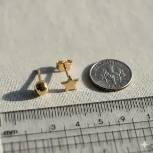 A macro image showing a pair of tiny 14k gold stud earrings next to a ruler or coin for an accurate size reference.