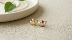 Side-by-side comparison of rose gold and yellow gold heart stud earrings on a neutral fabric background.
